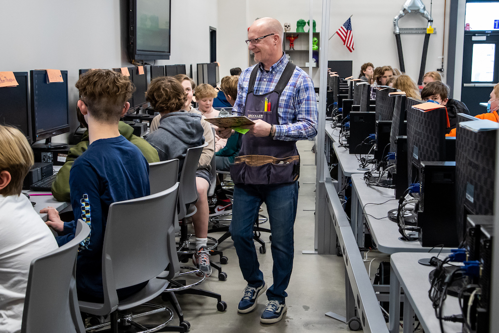 Male teacher wearing a black apron and carrying a clipboard walks behind students working at computers.